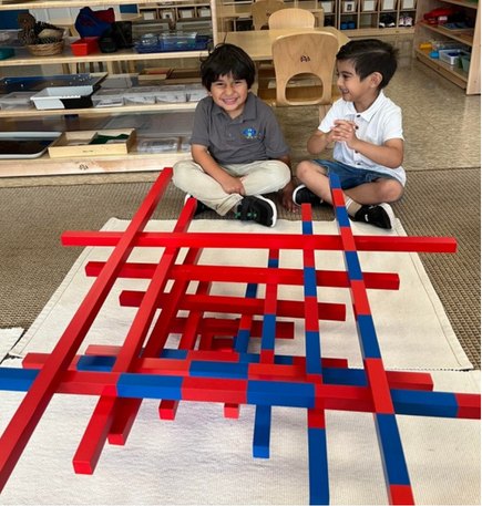 Two Seven Oaks students sit beside the Montessori red and blue number rods laid out on a rug